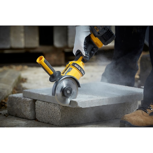 A DEWALT angle grinder cutting through a concrete block, operated by a person wearing gloves and work boots. Dust is visible around the cutting area.