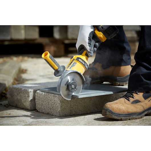 A person wearing work gloves and safety boots is using a yellow DEWALT power tool with a circular blade to cut a stone slab outdoors. Dust is visible as the tool operates.