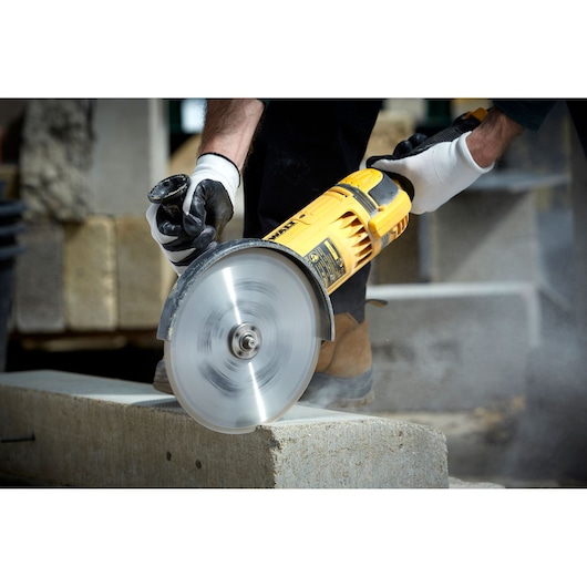 A person wearing gloves is using a yellow DEWALT power tool with a circular blade to cut through a concrete block outdoors.
