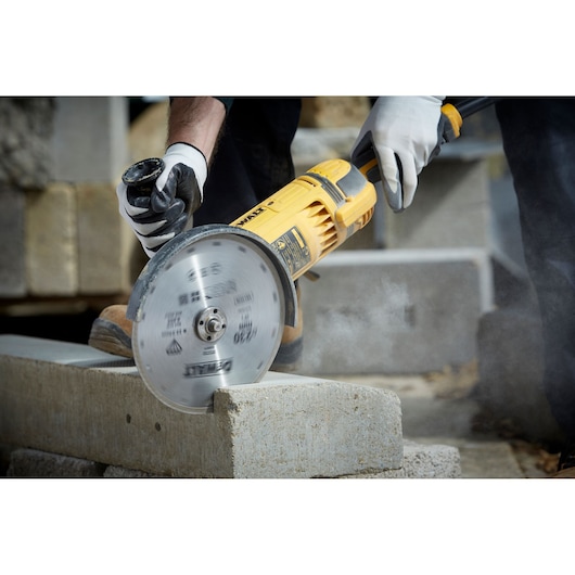 A close-up view of a person using a yellow DEWALT power tool with a circular blade to cut a concrete block. The person is wearing protective gloves.