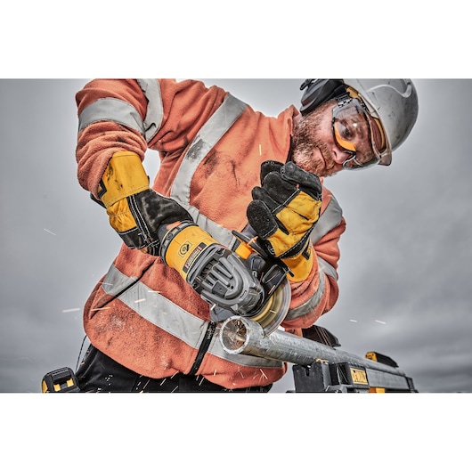 A person wearing an orange high-visibility jacket, yellow gloves, and a safety helmet is using a DEWALT angle grinder to cut a metal pipe. Sparks are visible around the cutting area. The person's face is obscured for privacy.