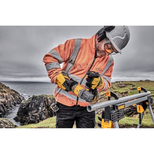 A person wearing safety gear, including a helmet, gloves, and an orange high-visibility jacket, is using a DEWALT power tool to grind or cut a metal pipe outdoors. The pipe is secured on a DEWALT stand, with rocky terrain and water visible in the background.