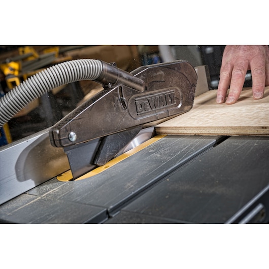 Close-up view of a DEWALT table saw in operation, with a dust extraction hose attached and a hand guiding a sheet of plywood through the blade.