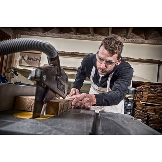 A person wearing an apron is using a DEWALT table saw to cut wooden planks in a workshop. The face of the person is blurred for anonymity. The workshop has shelves and stacks of wood in the background.