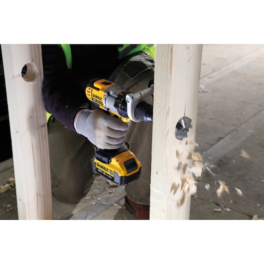 A person using a DEWALT cordless drill to bore a large hole through a wooden beam, with wood chips flying out on a construction site.