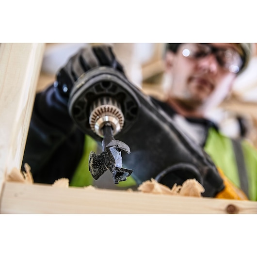 Close-up of a DEWALT DT4585 drill bit being used in a power drill to bore through wood, with wood shavings visible, and a person wearing gloves and safety gear in the background.