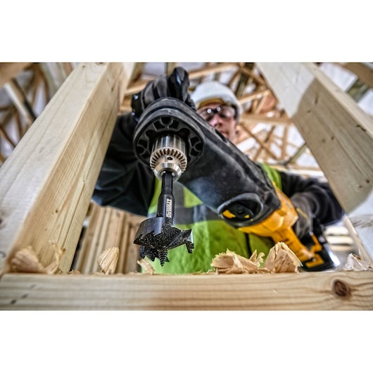 Close-up view of a DEWALT DT4585 drill bit in use, attached to a power drill, boring a hole through a wooden beam during construction. Wood shavings are visible around the area where the hole is being made.