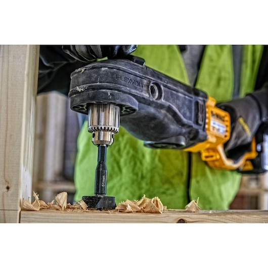 A close-up of a DEWALT power drill being used to drill a hole in a wooden beam, with wood shavings scattered around the drilled area.