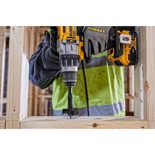 Close-up of a person using a DEWALT cordless drill with a special drill bit to make a hole in a wooden frame at a construction site. The person is wearing work gloves and a high-visibility jacket.