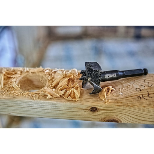 A DEWALT drill bit rests on a wooden plank next to wood shavings and a freshly drilled hole.