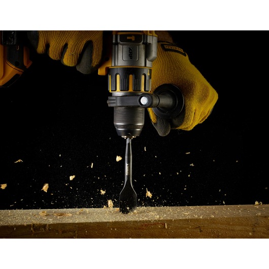 Close-up of a person wearing yellow gloves operating a DEWALT power drill with a spade bit, drilling into a piece of wood with wood chips flying.