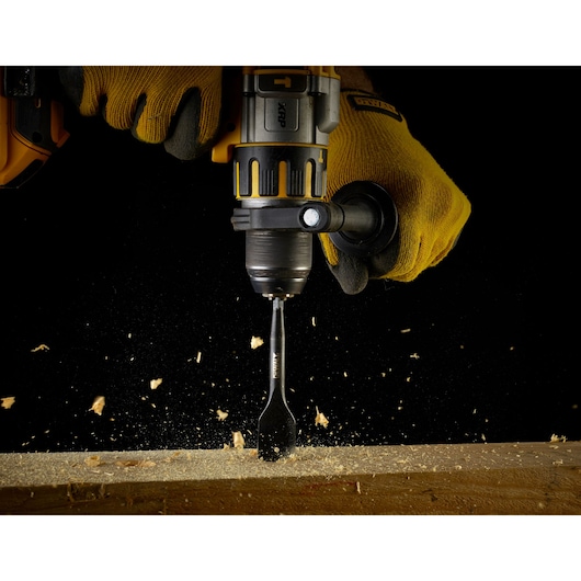 Close-up of a person wearing yellow work gloves using a DEWALT drill with a flat wood drill bit to bore into a piece of wood, with wood shavings flying.