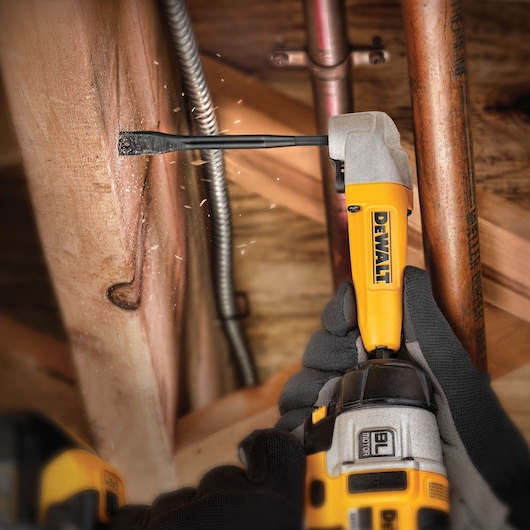 A DEWALT power drill fitted with a spade bit is being used to drill into a wooden beam, with wood chips flying. The drill is held by gloved hands, and copper pipes and metal conduit are visible in the background.