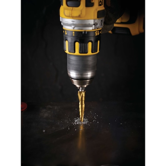 A DEWALT drill with a stepped drill bit is drilling into a dark metal surface, producing metal shavings.