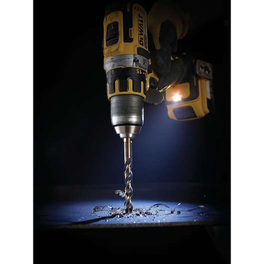 A DEWALT cordless drill with a spiral drill bit is being used to drill into a flat metal surface, with metal shavings scattered around the drilling area. The drill is illuminated by a built-in LED light.