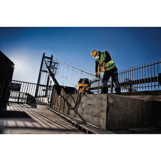 Worker uses DEWALT power tool and vacuum on concrete near a bridge, wearing safety gear and high-visibility vest.