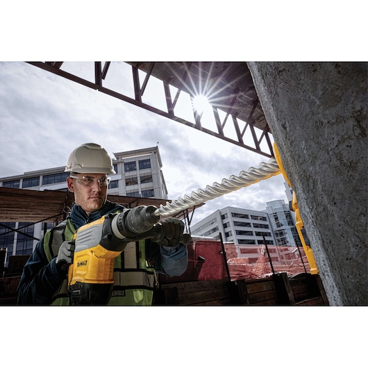 Worker uses a DEWALT drill with DT60816 bit on a concrete wall at a construction site, buildings in background.