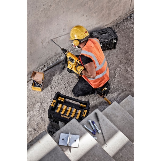 Worker in safety gear uses DEWALT drill near stairs; toolboxes and construction items are visible on the ground.