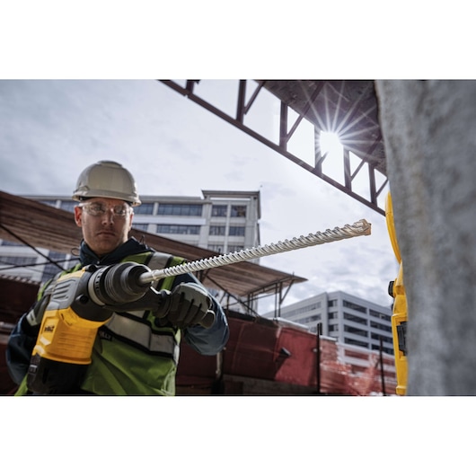 Worker uses a DEWALT rotary drill with long bit at a construction site with buildings in the background.