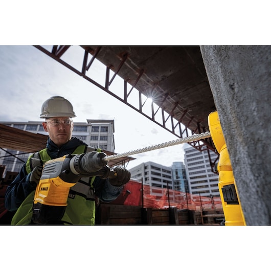 Worker using DEWALT drill with a long bit at a construction site, with urban buildings in the background.