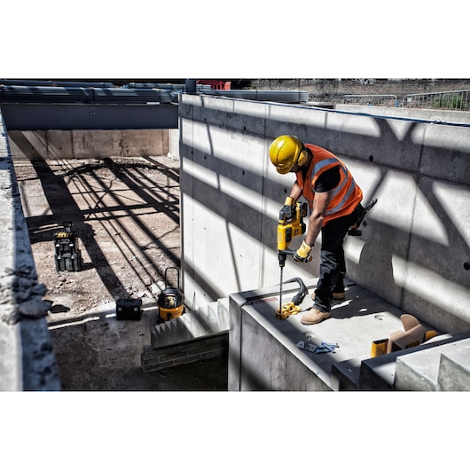 Worker uses a DEWALT drill on a concrete construction site, wearing safety gear and standing near scattered tools.