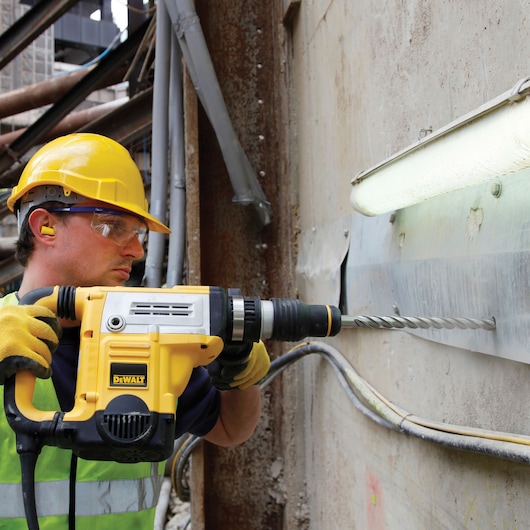 Worker uses a DEWALT hammer drill to bore holes in a concrete wall at a construction site, wearing safety gear.