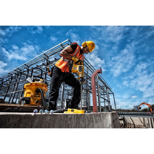 A construction worker wearing a yellow hard hat and orange safety vest uses a DEWALT power tool to drill into concrete at an outdoor construction site. Steel framework of a building and construction equipment are visible in the background.
