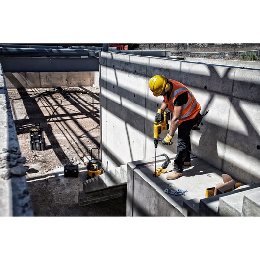 A construction worker wearing a yellow hard hat and orange safety vest is using a DEWALT power drill to work on a concrete structure at an outdoor construction site. Various tools and equipment are visible around the area.
