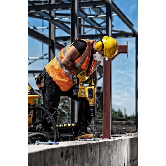 A construction worker wearing an orange safety vest, a yellow hard hat, ear protection, and a face mask is using a yellow DEWALT power drill to drill into a concrete surface at an outdoor construction site with steel framework in the background.