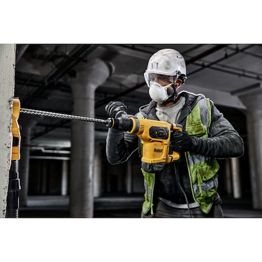 A construction worker wearing a safety helmet and reflective vest is using a DEWALT DT60827 rotary hammer drill to drill into a concrete pillar on a construction site.