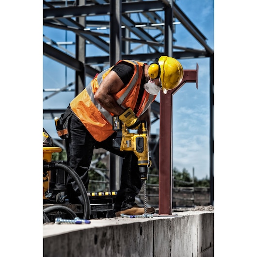 A construction worker wearing a hard hat, safety vest, ear protection, and a face mask uses a DEWALT cordless drill to drill into a concrete surface at a building site.