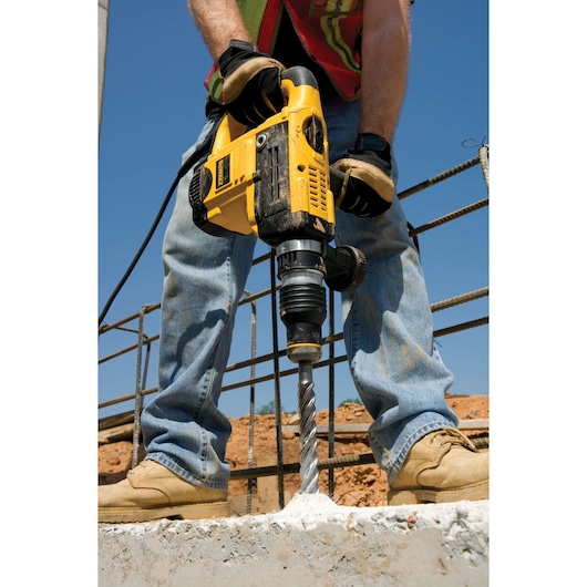 A person operating a yellow DEWALT rotary hammer drill to bore into concrete at a construction site. The drill is positioned vertically, and the person is wearing gloves, a safety vest, jeans, and work boots.