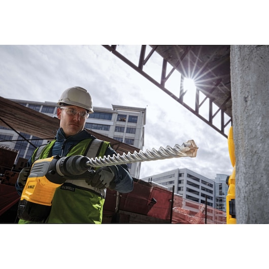Person holding a DEWALT DT60846 rotary hammer drill with spiral bit at a construction site, buildings in the background.