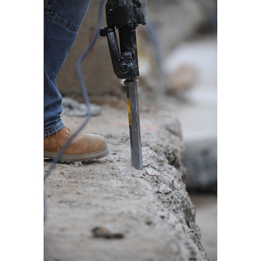 Close-up of a person using a power tool with a chisel bit to break concrete, wearing blue jeans and brown work boots.