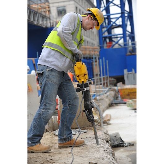 A construction worker wearing a yellow safety helmet, a high-visibility vest, and jeans is using a yellow DEWALT demolition hammer to break concrete on a construction site. The background shows other construction materials and a blue steel structure.