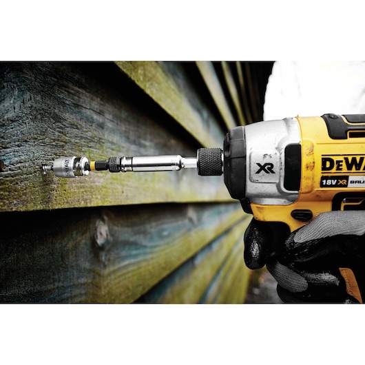 Close-up of a DEWALT cordless drill with a screwdriver bit in use, fastening a screw into a wooden wall. A gloved hand is holding the tool.