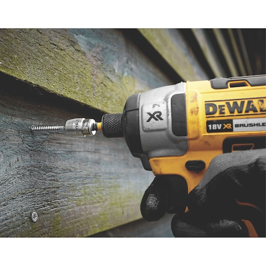 A close-up of a DEWALT 18V XR brushless power tool driving a screw into a wooden surface. The tool is held by a gloved hand, and the DEWALT logo is visible.