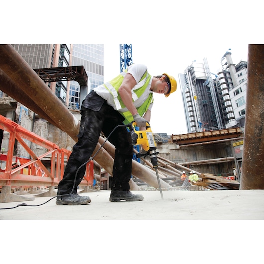 A construction worker wearing a yellow hard hat and reflective vest is using a DEWALT power tool to break concrete at a building site. Large metal pipes, safety barriers, and modern buildings are visible in the background.