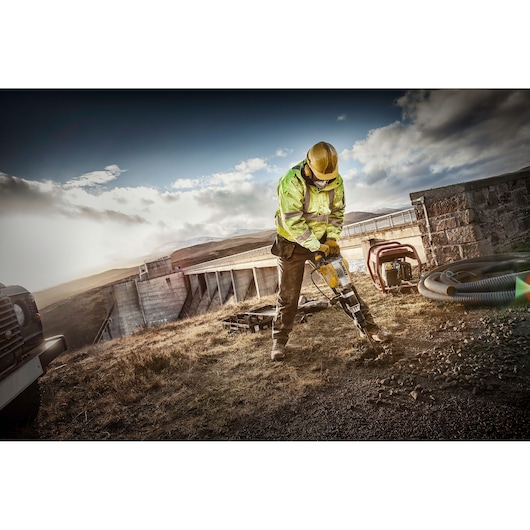 A construction worker in a high-visibility jacket and helmet using a DEWALT jackhammer to break up ground outdoors, near a large structure and generator, under a partly cloudy sky.