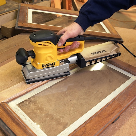 A DEWALT electric sander being used to sand the surface of a wooden window frame. The tool is held by a person and features a dust bag attachment.