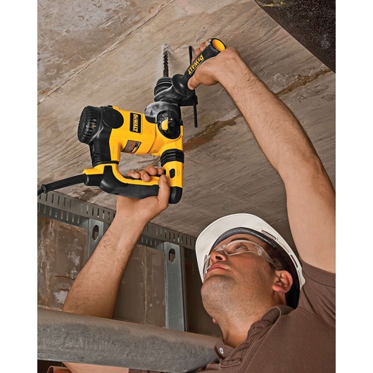 A person wearing a hard hat is using a DEWALT DT8911 rotary hammer drill to drill into a concrete ceiling. The drill is yellow and black, and the worker is holding it with both hands while reaching upward.