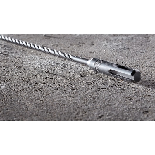 A close-up image of a DEWALT DT8914 SDS-plus drill bit lying on a rough concrete surface. The metal bit features a spiral design and is designed for masonry and concrete drilling.