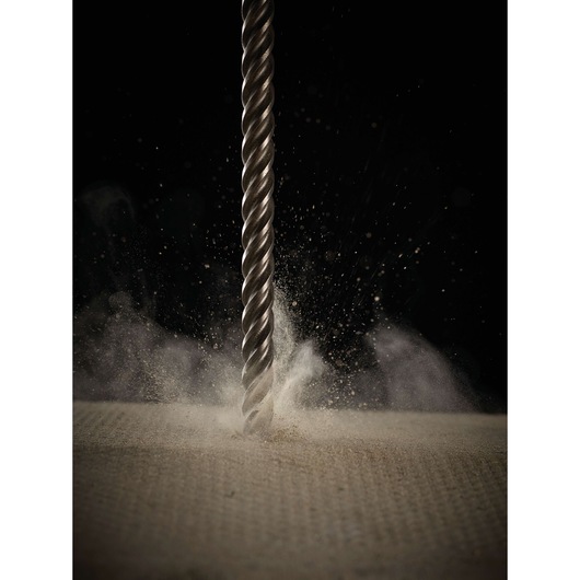 Close-up of a metallic drill bit penetrating a surface, with dust and particles being scattered, against a dark background.
