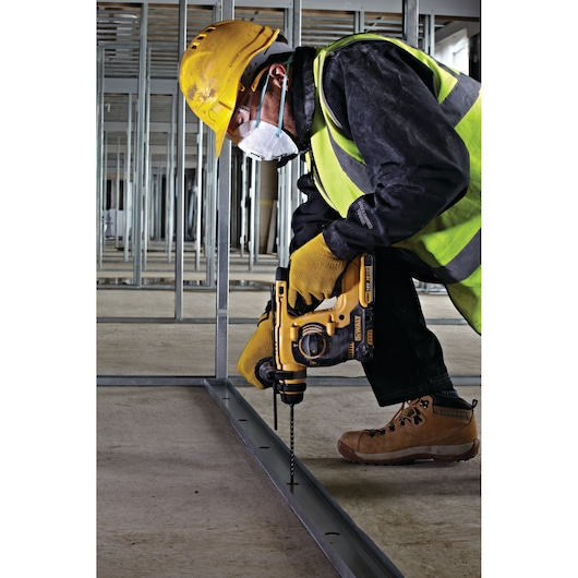 A construction worker wearing a yellow hard hat, safety vest, gloves, and a protective mask is using a DEWALT power drill to drill into a metal frame on the floor in an unfinished building structure.