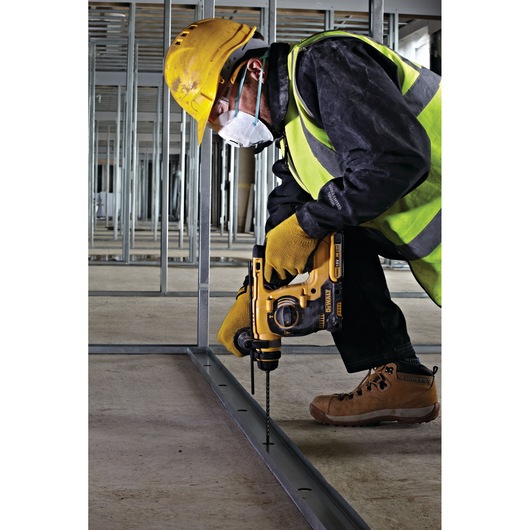A construction worker wearing a yellow helmet, safety gloves, a high-visibility vest, protective boots, and a mask is using a yellow DEWALT rotary hammer drill to drill into a metal track on a concrete floor at a building site framed with steel studs.