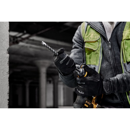 A person wearing gloves and a green safety vest is holding a DEWALT power drill with a spiral drill bit attached, in an industrial setting.