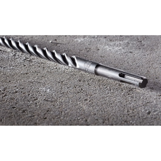 A close-up image of a DEWALT DT8938 rotary hammer drill bit with a spiral design, lying on a rough concrete surface.