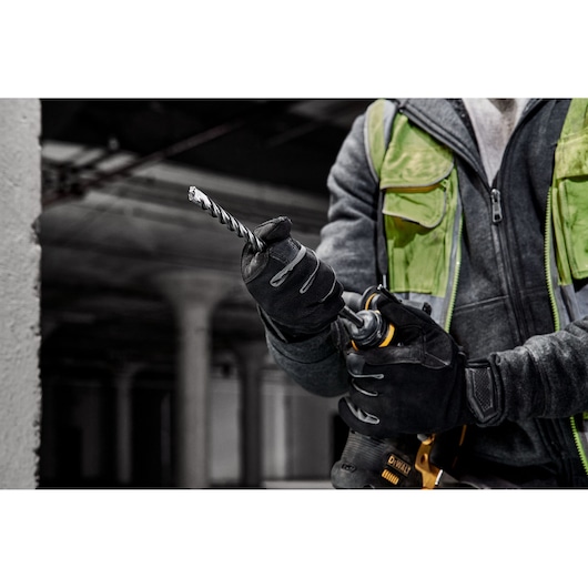 A person wearing work gloves and a green safety vest is holding a DEWALT power drill with a spiral drill bit, preparing for use at a construction site.