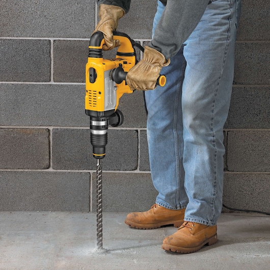 A person wearing gloves, jeans, and work boots is using a yellow DEWALT hammer drill to drill into a concrete floor, with a cinder block wall in the background.