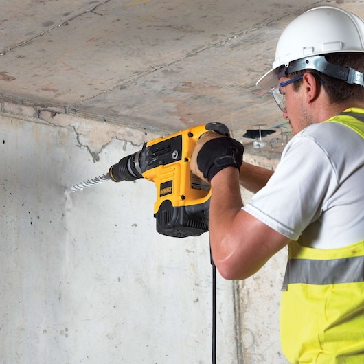 A person wearing a white hard hat, safety vest, gloves, and protective glasses is using a yellow DEWALT rotary hammer drill to drill into a concrete wall.